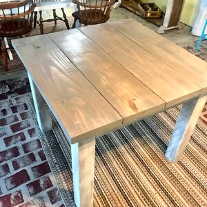 May include: A square wooden table with a light gray stain finish. The table has four legs and a simple design. The table is sitting on a striped rug.