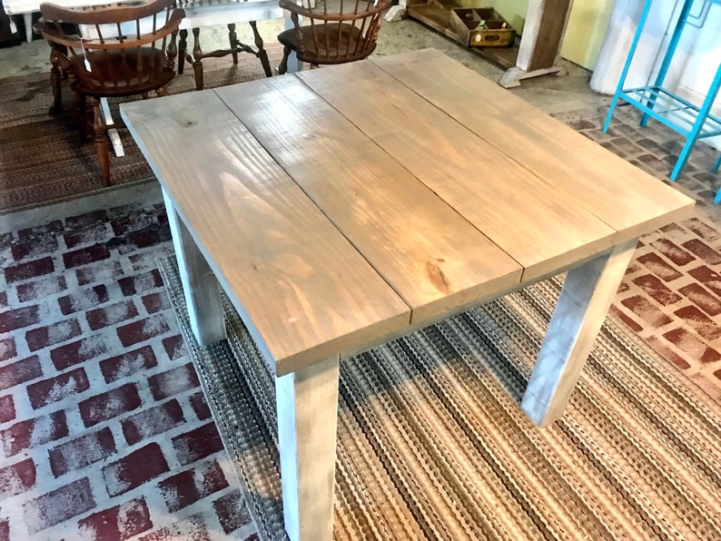 May include: A square wooden table with a light gray stain finish. The table has four legs and a simple design. The table is sitting on a striped rug.