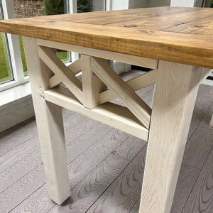 May include: A wooden table with a light brown top and white legs. The table has a rectangular top and a cross-braced design on the sides. The table is made of wood and has a rustic appearance.