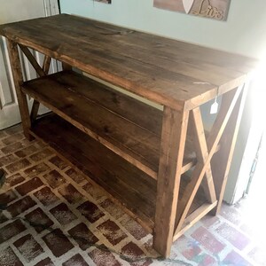 May include: A rectangular wooden console table with a dark brown stained finish. The piece features a flat top surface, one lower shelf, and side panels constructed with a prominent X-shaped cross-brace design. The structure is made of planks.
