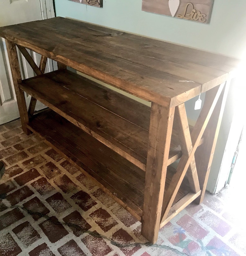 May include: A rectangular wooden console table with a dark brown stained finish. The piece features a flat top surface, one lower shelf, and side panels constructed with a prominent X-shaped cross-brace design. The structure is made of planks.