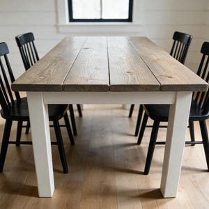 May include: A rectangular dining table with a natural wood grain top and four white legs, accompanied by four black spindle-back chairs. The table features a plank-style surface with visible wood knots and a smooth, matte white finish on the base.