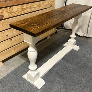 May include: A rectangular console table featuring a dark brown stained wooden top with visible wood grain and knots. The table is supported by two white painted pedestal bases, each consisting of a turned column post on a rectangular base.