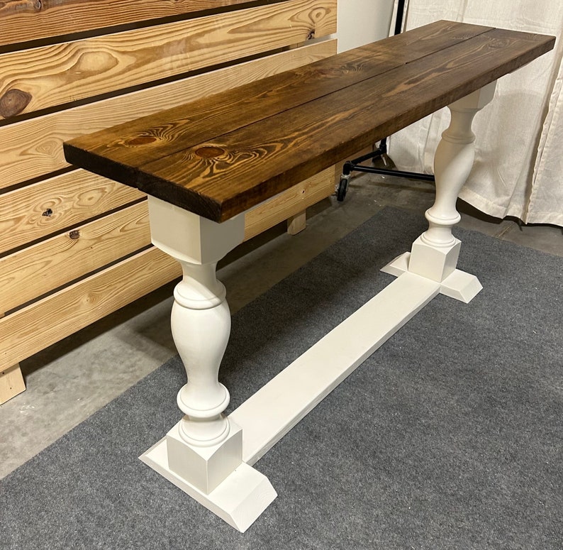 May include: A rectangular console table featuring a dark brown stained wooden top with visible wood grain and knots. The table is supported by two white painted pedestal bases, each consisting of a turned column post on a rectangular base.