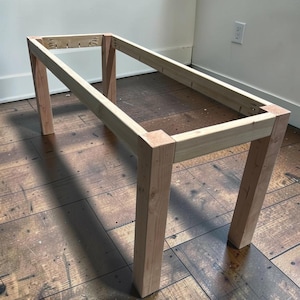 May include: A rectangular wooden table frame, constructed from light-colored lumber. The frame features four sturdy legs and a top structure, showcasing the basic form of a table. The unfinished wood displays natural grain patterns, suggesting a DIY or handcrafted project.