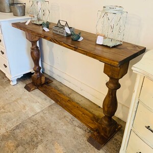 May include: A long, narrow wooden console table with a dark brown stained finish. The table features a rectangular top supported by two thick, turned pedestal legs connected by a horizontal base stretcher that runs along the floor.