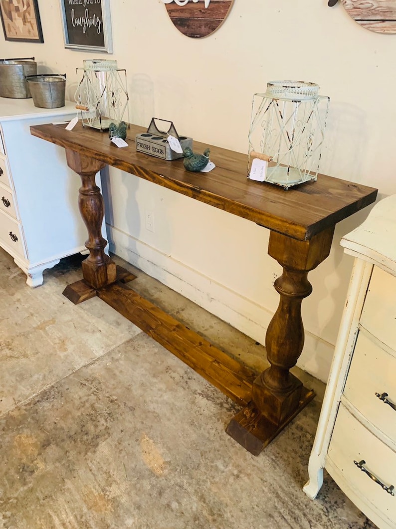 May include: A long, narrow wooden console table with a dark brown stained finish. The table features a rectangular top supported by two thick, turned pedestal legs connected by a horizontal base stretcher that runs along the floor.