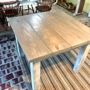 May include: A square wooden table with a gray stain finish and white painted legs. The table is sitting on a rug with a brick pattern.