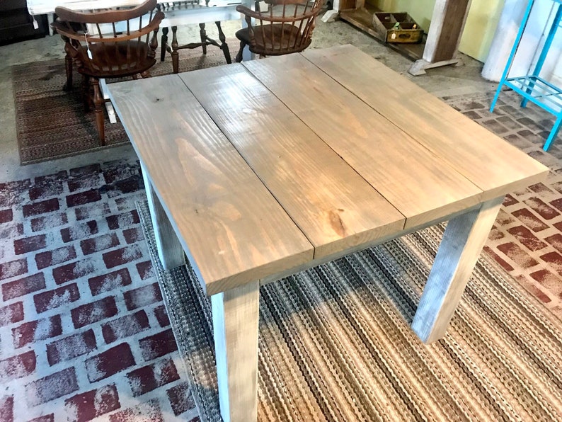May include: A square wooden table with a gray stain finish and white painted legs. The table is sitting on a rug with a brick pattern.