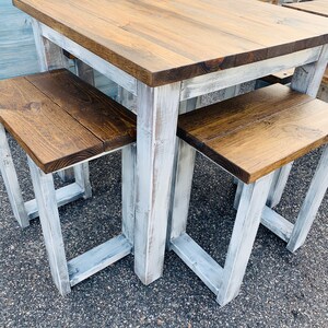K&ouml;nnte beinhalten: Ein quadratischer Holztisch mit einer dunkelbraun gebeizten Tischplatte und wei&szlig; lackierten Beinen im Used-Look. Zwei passende Holzb&auml;nke mit dunkelbraun gebeizten Sitzfl&auml;chen und wei&szlig; lackierten Beinen im Used-Look stehen unter dem Tisch.