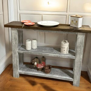 May include: A whitewashed wooden console table with two shelves. The table has a distressed finish and is decorated with various items, including a red rooster figurine, a white ceramic jar, and a white ceramic box with a floral design. The table is sitting on a wooden floor.