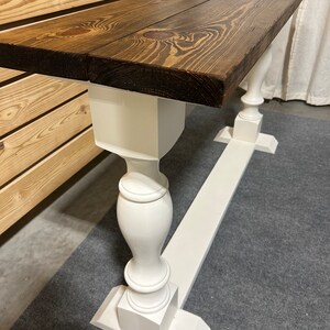 May include: A long, narrow console table featuring a thick, dark brown stained wooden top with visible wood grain and knots. The table is supported by two white painted pedestal-style legs connected by a long, flat white base board.