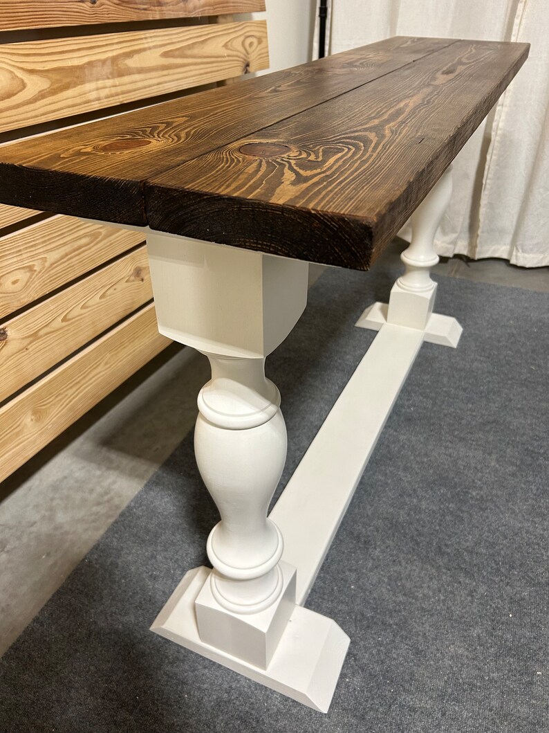 May include: A long, narrow console table featuring a thick, dark brown stained wooden top with visible wood grain and knots. The table is supported by two white painted pedestal-style legs connected by a long, flat white base board.
