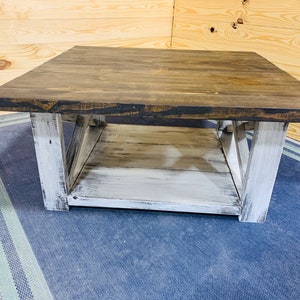 May include: A square coffee table featuring a dark brown stained wooden top and a lower shelf. The base consists of four thick, white distressed wooden legs connected by X-shaped side supports, creating a sturdy frame with a two-tone finish.