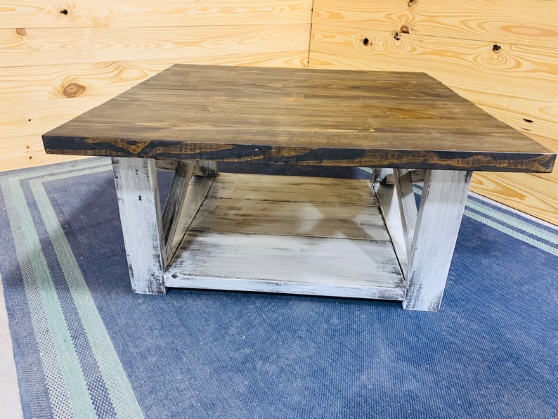 May include: A square coffee table featuring a dark brown stained wooden top and a lower shelf. The base consists of four thick, white distressed wooden legs connected by X-shaped side supports, creating a sturdy frame with a two-tone finish.