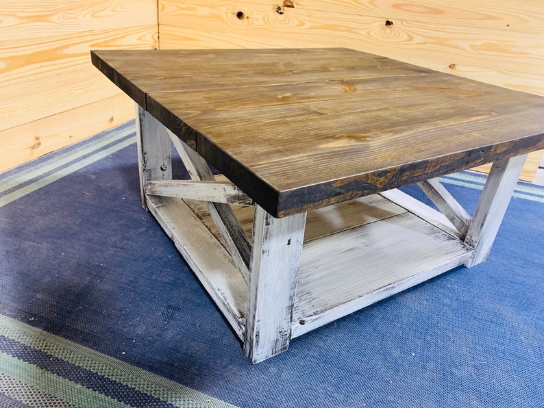 May include: A square coffee table featuring a dark brown stained wooden top and a lower shelf. The base is constructed with white painted wooden legs and cross-bracing supports that have a distressed, weathered finish on the edges.