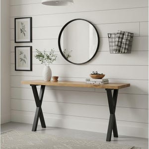 May include: A rectangular wooden console table with a natural light brown finish and black metal X-shaped legs. The table surface holds a white vase with greenery, a small wooden bowl, and a stack of books. A round black-framed mirror hangs above.