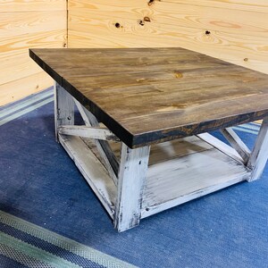 May include: A square coffee table featuring a dark brown stained wooden top and a distressed white painted base. The base includes four sturdy legs connected by X-shaped support braces on each side and a lower shelf for additional storage space.