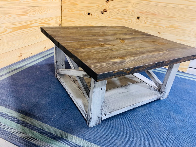 May include: A square coffee table featuring a dark brown stained wooden top and a distressed white painted base. The base includes four sturdy legs connected by X-shaped support braces on each side and a lower shelf for additional storage space.