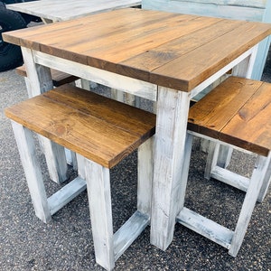 K&ouml;nnte beinhalten: Ein quadratischer Holztisch mit dunkelbraun gebeizter Tischplatte und einem wei&szlig; gestrichenen Untergestell im Used-Look, begleitet von zwei passenden r&uuml;ckenlehnenlosen B&auml;nken mit dunkelbraunen Sitzen und wei&szlig; lackierten Holzbeinen.