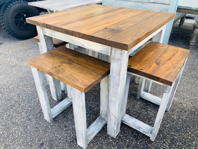 K&ouml;nnte beinhalten: Ein quadratischer Holztisch mit dunkelbraun gebeizter Tischplatte und einem wei&szlig; gestrichenen Untergestell im Used-Look, begleitet von zwei passenden r&uuml;ckenlehnenlosen B&auml;nken mit dunkelbraunen Sitzen und wei&szlig; lackierten Holzbeinen.