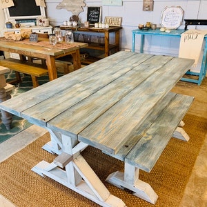 May include: A large, rustic farmhouse-style dining table with a matching bench. The tabletop is made of light blue-gray stained wood planks. The base and legs are painted white, creating a contrasting two-tone effect. The table sits on a woven jute rug.