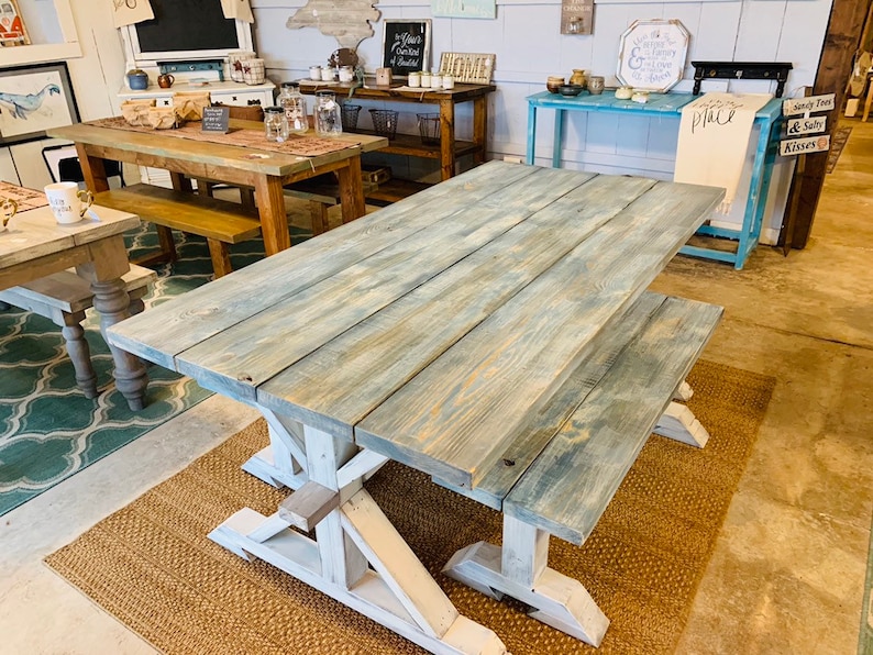 May include: A large, rustic farmhouse-style dining table with a matching bench. The tabletop is made of light blue-gray stained wood planks. The base and legs are painted white, creating a contrasting two-tone effect. The table sits on a woven jute rug.