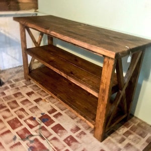 May include: A rectangular wooden console table with a dark brown stained finish. The piece features a flat top surface, one lower shelf, and side supports constructed with a prominent X-shaped cross-brace design on both the left and right ends.