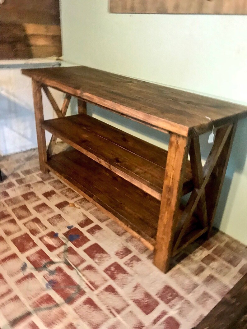 May include: A rectangular wooden console table with a dark brown stained finish. The piece features a flat top surface, one lower shelf, and side supports constructed with a prominent X-shaped cross-brace design on both the left and right ends.