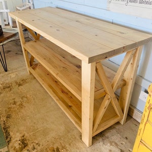 May include: A rectangular wooden console table featuring a natural light brown finish, a flat top surface, and one lower shelf. The structure is supported by four thick legs connected by decorative X-shaped cross-bracing on both side panels.