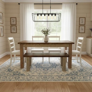May include: A rectangular wooden dining table with a dark brown top and lighter legs, accompanied by a matching wooden bench, four white ladder-back chairs, and a black metal rectangular chandelier with six exposed filament light bulbs.