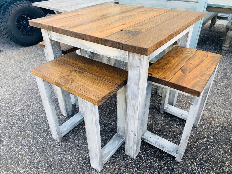 K&ouml;nnte beinhalten: Ein quadratischer Holztisch mit einer mittelbraun gebeizten Tischplatte und einem wei&szlig; gestrichenen Untergestell im Used-Look, dazu zwei passende rechteckige B&auml;nke mit braun gebeizten Sitzfl&auml;chen und wei&szlig; gestrichenen Beinen im Used-Look.