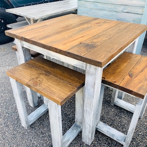 K&ouml;nnte beinhalten: Ein quadratischer Holztisch mit einer dunkelbraun gebeizten Tischplatte und einem wei&szlig; gestrichenen Gestell im Used-Look, begleitet von zwei passenden r&uuml;ckenlehnenlosen Hockern mit dunkelbraunen Holzsitzen und wei&szlig;em Gestell.