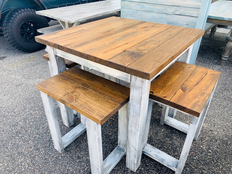 K&ouml;nnte beinhalten: Ein quadratischer Holztisch mit einer dunkelbraun gebeizten Tischplatte und einem wei&szlig; gestrichenen Gestell im Used-Look, begleitet von zwei passenden r&uuml;ckenlehnenlosen Hockern mit dunkelbraunen Holzsitzen und wei&szlig;em Gestell.