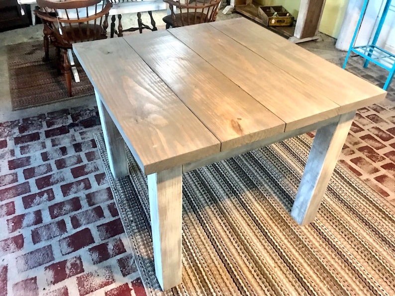 May include: A square wooden table with a light gray stain finish. The table has four legs and is sitting on a woven rug.