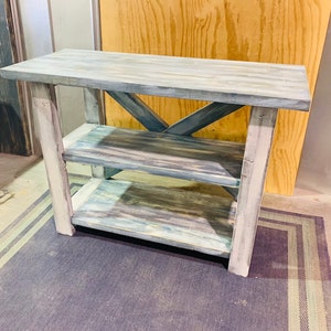 May include: A rectangular wooden console table featuring a distressed finish with shades of blue, gray, and white. The piece includes a flat top surface, two lower shelves, and a structural X-shaped support brace on the back side between the legs.