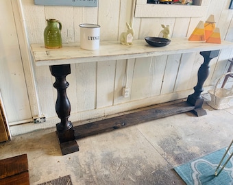 May include: A long console table featuring a distressed white rectangular top supported by two dark brown turned wooden legs connected by a horizontal base beam. The table surface holds a green ceramic pitcher, a white utensil crock, two small rabbit figurines, a black bowl, and three triangular candy corn decorations.
