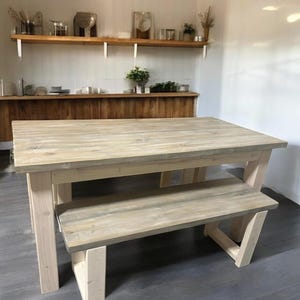 May include: A light-colored wooden dining table with a matching bench. The table and bench have a rustic, farmhouse style. The table is rectangular, and the bench fits neatly underneath. The wood has a natural, weathered appearance, and the legs are a lighter shade.
