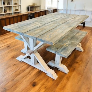 May include: A rustic wooden dining table and bench set. The tabletop and bench have a weathered, gray-blue finish, while the trestle-style base is painted white. The set is placed on a hardwood floor.