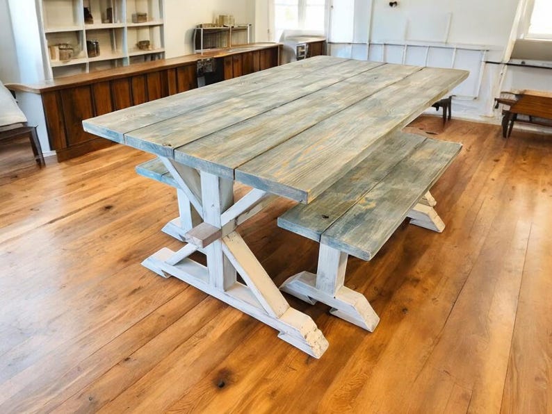 May include: A rustic wooden dining table and bench set. The tabletop and bench have a weathered, gray-blue finish, while the trestle-style base is painted white. The set is placed on a hardwood floor.
