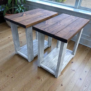 May include: Two rectangular side tables featuring dark brown stained wooden tops and distressed white painted wooden legs and lower shelves. Each table has a sturdy four-legged frame with a flat surface and a matching shelf positioned near the floor.