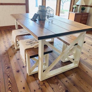 May include: A rustic dining table with a dark stained top and white base. Two matching benches are tucked underneath. The table is made of wood and has a farmhouse style. The benches have cream-colored throws with black text.