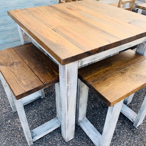 K&ouml;nnte beinhalten: Ein quadratischer Holztisch mit einer mittelbraun gebeizten Tischplatte und einem wei&szlig; gestrichenen, abgenutzten Untergestell, dazu zwei passende B&auml;nke mit braunen Sitzen und wei&szlig;en Beinen, mit rustikalem Finish und sichtbarer Holzmaserung.