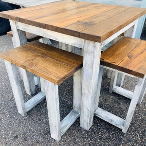K&ouml;nnte beinhalten: Ein quadratischer Holztisch mit dunkelbraun gebeizter Platte und einem wei&szlig; gestrichenen Gestell im Used-Look, dazu zwei passende B&auml;nke mit dunkelbraun gebeizten Sitzfl&auml;chen und wei&szlig; gestrichenen Beinen im Used-Look, alle mit stabilem Rahmen.
