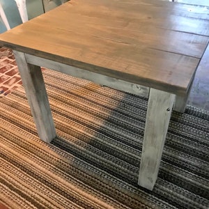 May include: A square wooden table with a distressed white paint finish. The table top is a light brown wood with a natural wood grain. The table is sitting on a striped rug.