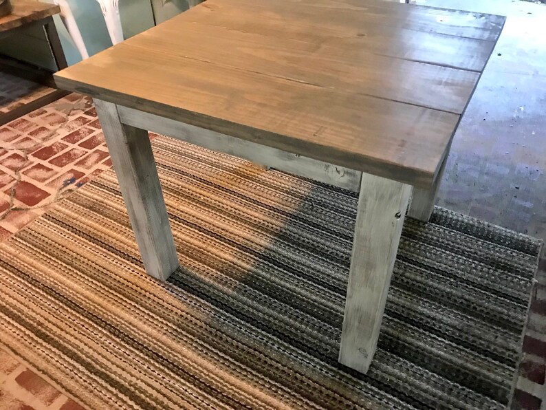 May include: A square wooden table with a distressed white paint finish. The table top is a light brown wood with a natural wood grain. The table is sitting on a striped rug.