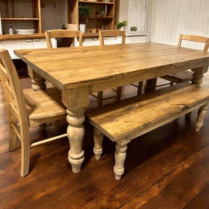 May include: A rectangular wooden dining table with a natural light brown finish, featuring thick, turned legs. The set includes two matching wooden chairs with horizontal back slats and a long wooden bench with matching turned legs.