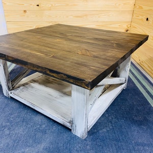 May include: A square coffee table featuring a dark brown stained wooden top and a white distressed painted base. The base includes four sturdy legs connected by X-shaped side supports and a lower shelf for storage, showcasing a two-tone finish.