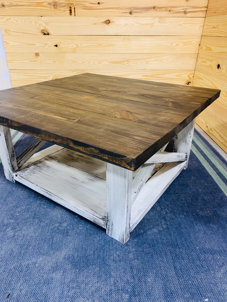 May include: A square coffee table featuring a dark brown stained wooden top and a white distressed painted base. The base includes four sturdy legs connected by X-shaped side supports and a lower shelf for storage, showcasing a two-tone finish.