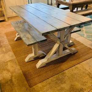 May include: A rustic farmhouse-style dining table and bench set. The table features a distressed gray-blue tabletop and white-painted trestle-style legs. The bench mirrors the table's design. The set is placed on a woven brown rug.